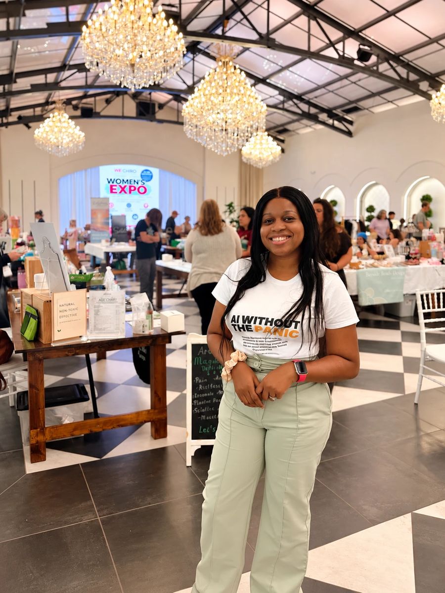 Dr. Gbemisola Adetayo smiles in the Women’s Expo hall, wearing an AI Without the Panic T-shirt, with vendor tables, chandeliers and a Women’s Expo screen behind her
