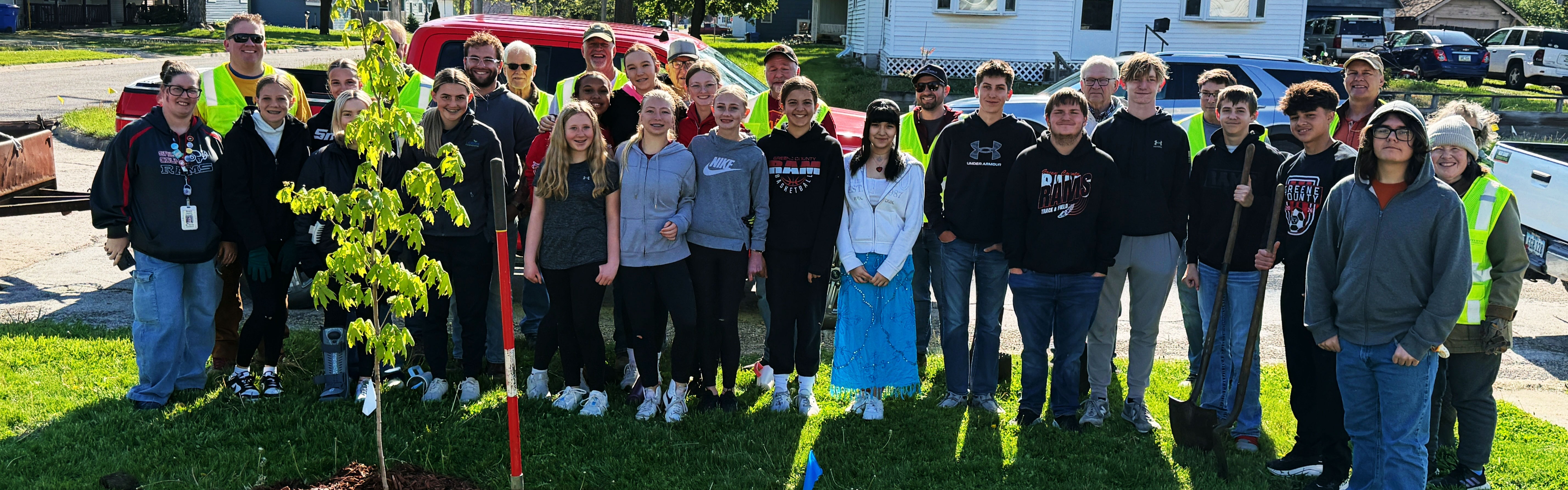 Community volunteers and students of mixed ages standing together outdoors on a sunny day beside a newly planted tree.