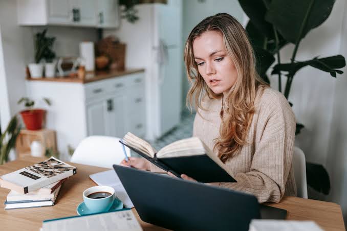A woman sits at a sunlit kitchen table with an open laptop, reading from a notebook she is holding and writing in it with a pencil.