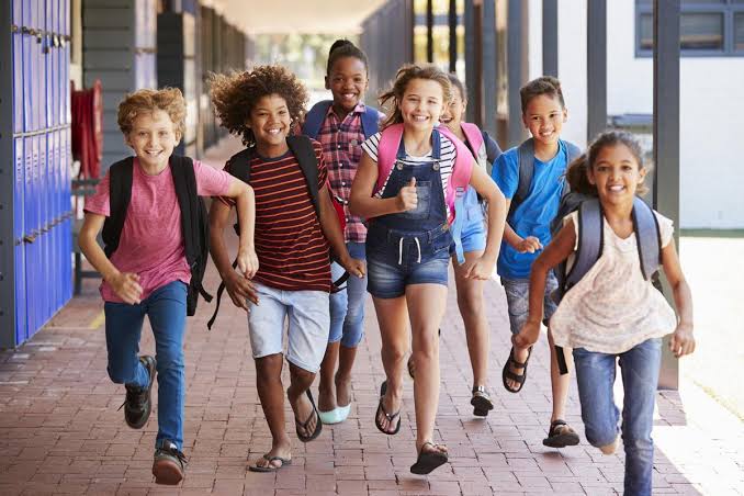 Six school-age children with backpacks running and laughing along a school corridor