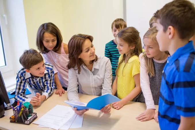 A teacher seated at a desk holding an open folder, talking with a group of children gathered around her