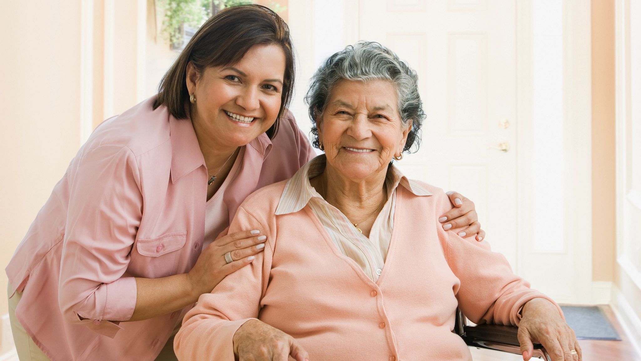 A woman standing with her arm around an older woman seated in a bright hallway, both smiling