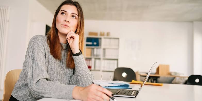 A woman sitting at a desk with a laptop and papers, resting her chin on her hand and looking up in thought