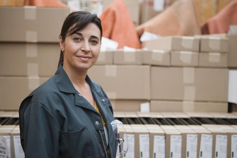 A woman in a work jacket holding safety glasses, standing among stacked cartons in a storeroom
